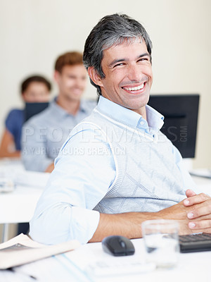 Buy stock photo Portrait of a positive-looking office worker sitting at his workstation with colleagues sitting behind him