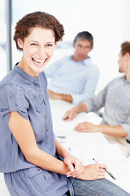 Buy stock photo Portrait of a laughing female office worker sitting on her desk with colleagues sitting behind her