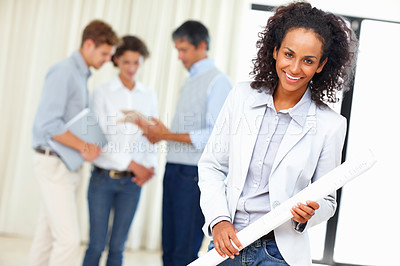 Buy stock photo Young business woman holding blueprint with colleagues discussing in background
