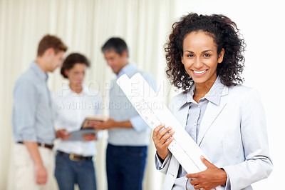Buy stock photo Young business woman holding blueprints with colleagues discussing in background