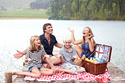 Buy stock photo Happy family, picnic and relax with rain at lake outdoor to celebrate, carefree or people laugh on water jetty. Mother, father or kids at river with food on vacation with hand gesture for wet weather