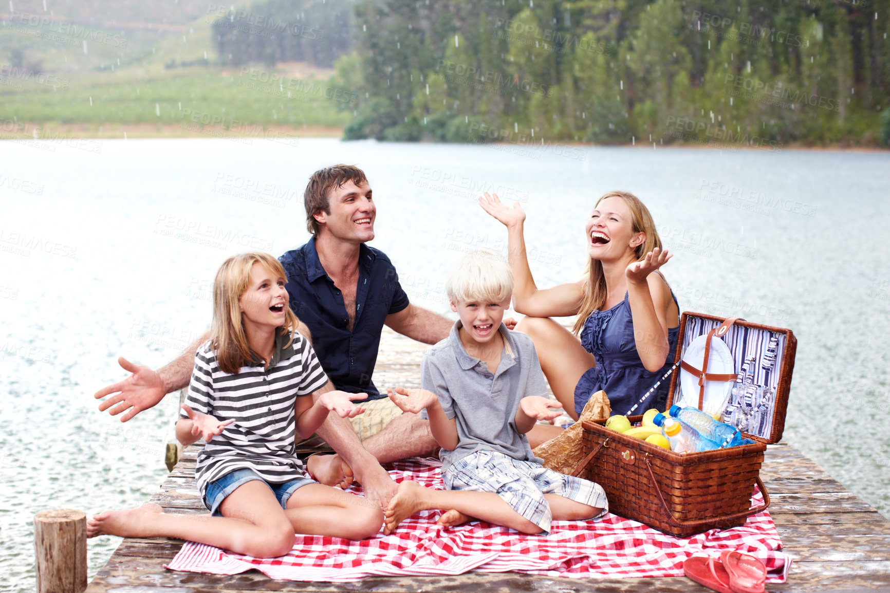 Buy stock photo Happy family, picnic and relax with rain at lake outdoor to celebrate, carefree or people laugh on water jetty. Mother, father or kids at river with food on vacation with hand gesture for wet weather