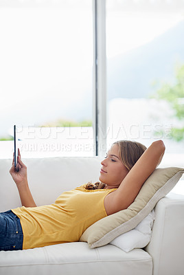 Buy stock photo Shot of a young woman using a digital tablet while lying on a sofa at home