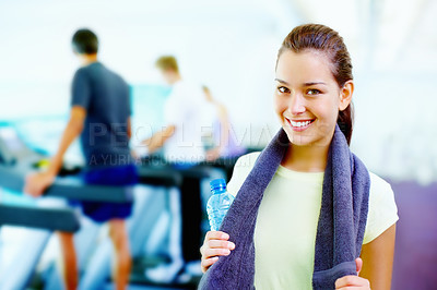 Buy stock photo Portrait of young smiling woman holding water bottle and towel with people working out in background