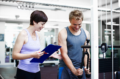 Buy stock photo Cropped shot of a handsome young man exercising in the gym while his personal trainer takes notes