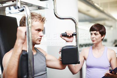 Buy stock photo Cropped shot of a handsome young man using an exercise machine while his personal trainer looks on