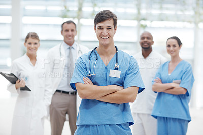 Buy stock photo Young male doctor standing smiling with his arms folded and his colleagues in the background