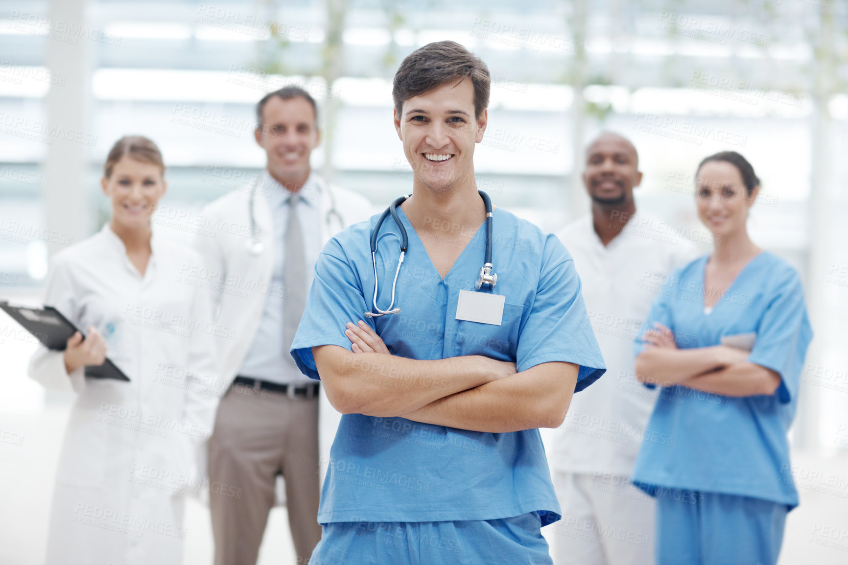 Buy stock photo Young male doctor standing smiling with his arms folded and his colleagues in the background