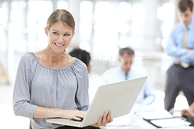 Buy stock photo A happy young woman using her laptop in the office with her colleagues behind her