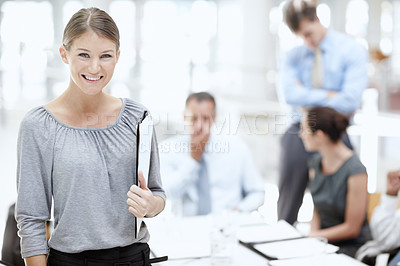 Buy stock photo A young woman standing with a notebook in the office with her colleagues behind her