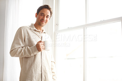 Buy stock photo Portrait of handsome man drinking coffee by his window in his pajamas in the morning. A young male wearing pyjamas and relaxing in his bright bedroom on the weekend. Enjoying his morning routine