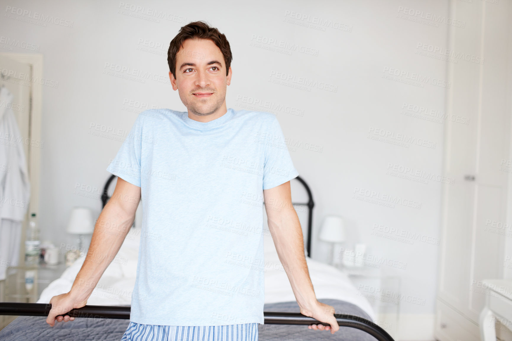 Buy stock photo A man standing by his bed dressed in comfortable pajamas or lounge wear and looking away. A young brunette male relaxing in his modern bright bedroom at home on the weekend