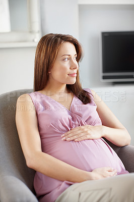 Buy stock photo Pretty young pregnant woman looking away while she sits in her living room as she holds her belly
