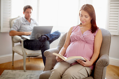 Buy stock photo Calm young pregnant woman reading in her living room with her boyfriend in the background