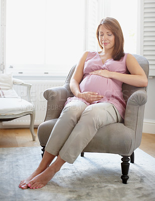 Buy stock photo Young pregnant woman holding her stomach while she sits in her living room at home