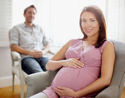 Buy stock photo Portrait of a beautiful pregnant woman holding her stomach while her husband sits in the background