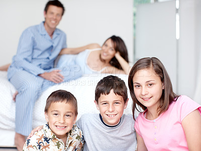 Buy stock photo Portrait of three siblings smiling as their parents sit blurred in the background