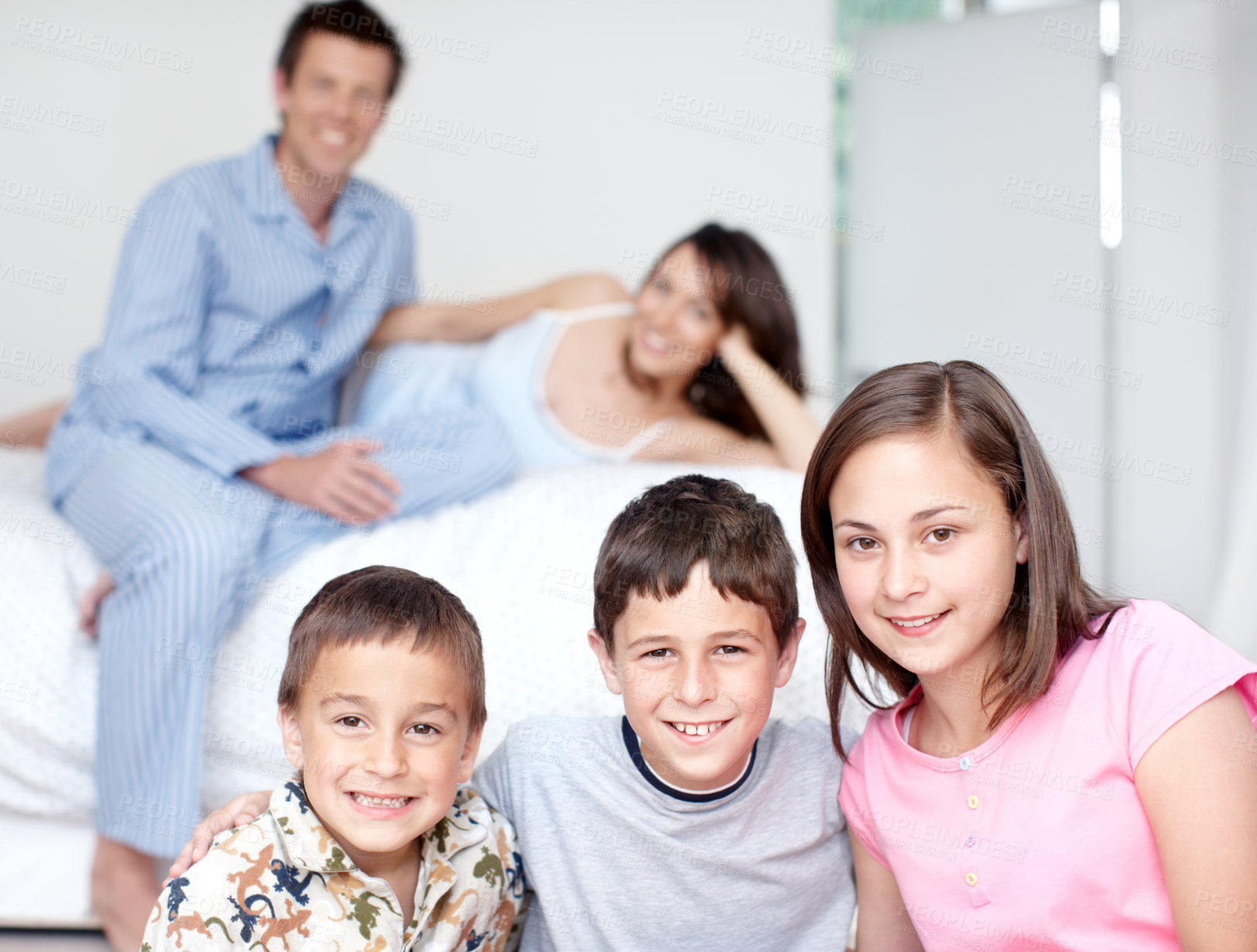 Buy stock photo Portrait of three siblings smiling as their parents sit blurred in the background