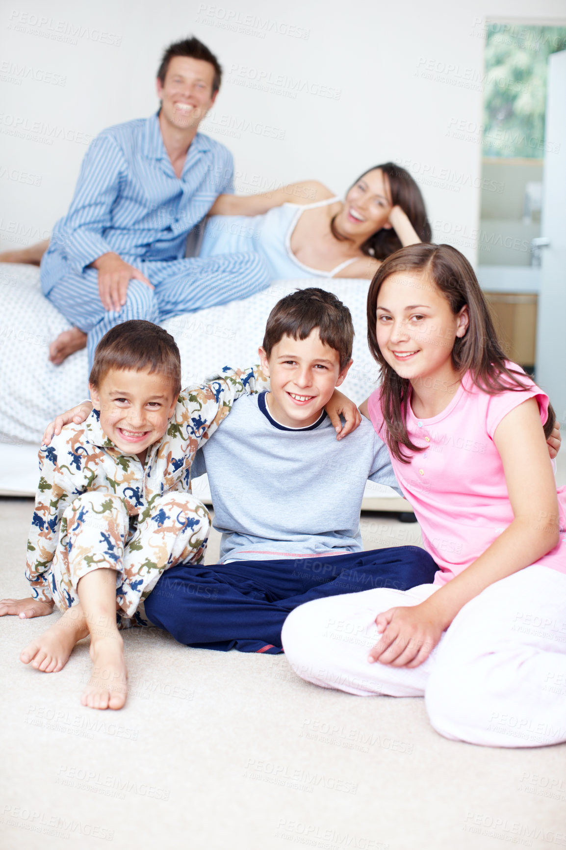 Buy stock photo Portrait of three siblings embracing as their parents sit blurred in the background