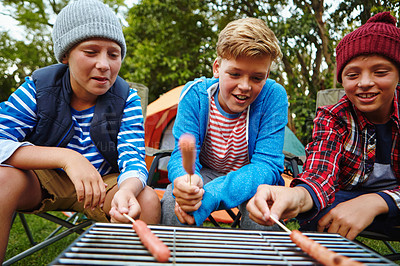 Buy stock photo Cropped shot of a three boys roasting hotdogs on a grill while camping