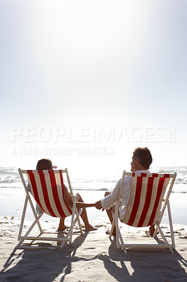 Buy stock photo Rearview of a happy couple sitting on deck-chairs on the beach and holding hands