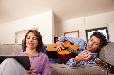 Buy stock photo Shot of a woman using a digital tablet while her husband plays the guitar