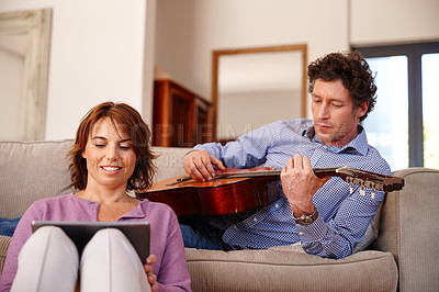 Buy stock photo Shot of a woman using a digital tablet while her husband plays the guitar