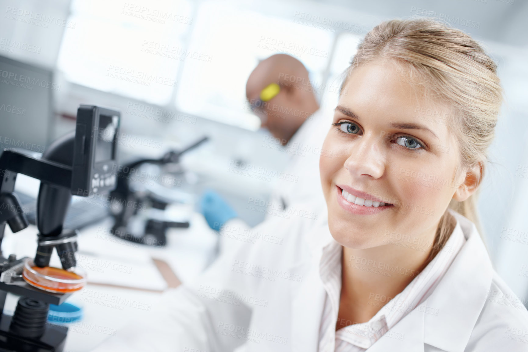 Buy stock photo Closeup portrait of a gorgeous female scientist in front of a microscope with a male colleague in the background
