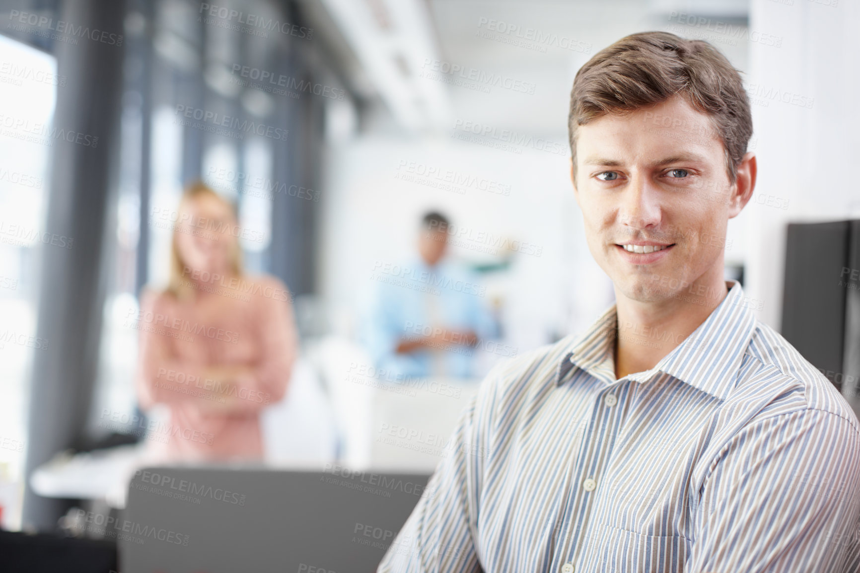 Buy stock photo Portrait of a young male designer standing at his desk with colleagues in the background in an office at work. Business professional ready for work. Businessman working at a startup company