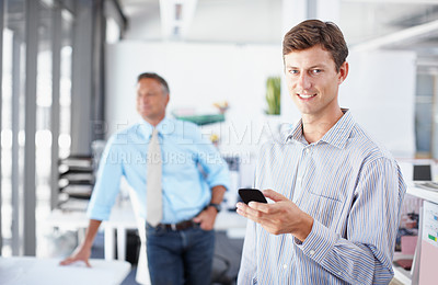 Buy stock photo A young professional holding a smartphone while a colleague stands in the background