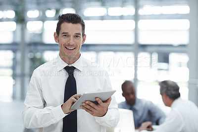 Buy stock photo A handsome businessman holding a touchpad, smiling and looking at the camera with colleagues working in the background