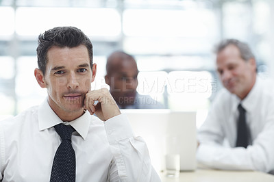 Buy stock photo A handsome businessman sitting with his chin rested on his hand looking at the camera with two colleagues in the background working on a laptop