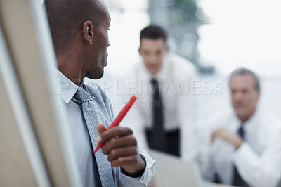 Buy stock photo African businessman standing at flipchart with a marking pen looking over his shoulder at colleagues in the background