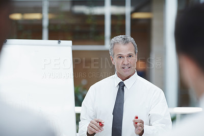 Buy stock photo A senior businessman standing besides a flipchart with a marker in hand and talks to his colleagues in the foreground