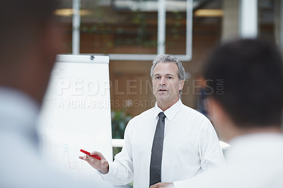 Buy stock photo A senior businessman standing infront of a flipchart with a marker in his hand and talks to his colleagues in the foreground
