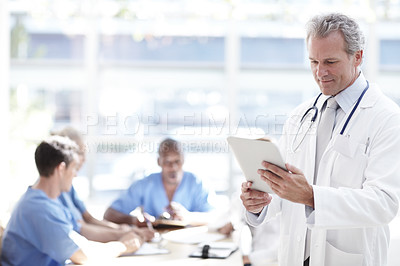 Buy stock photo A senior doctor holding a touchpad with colleagues sitting in the background