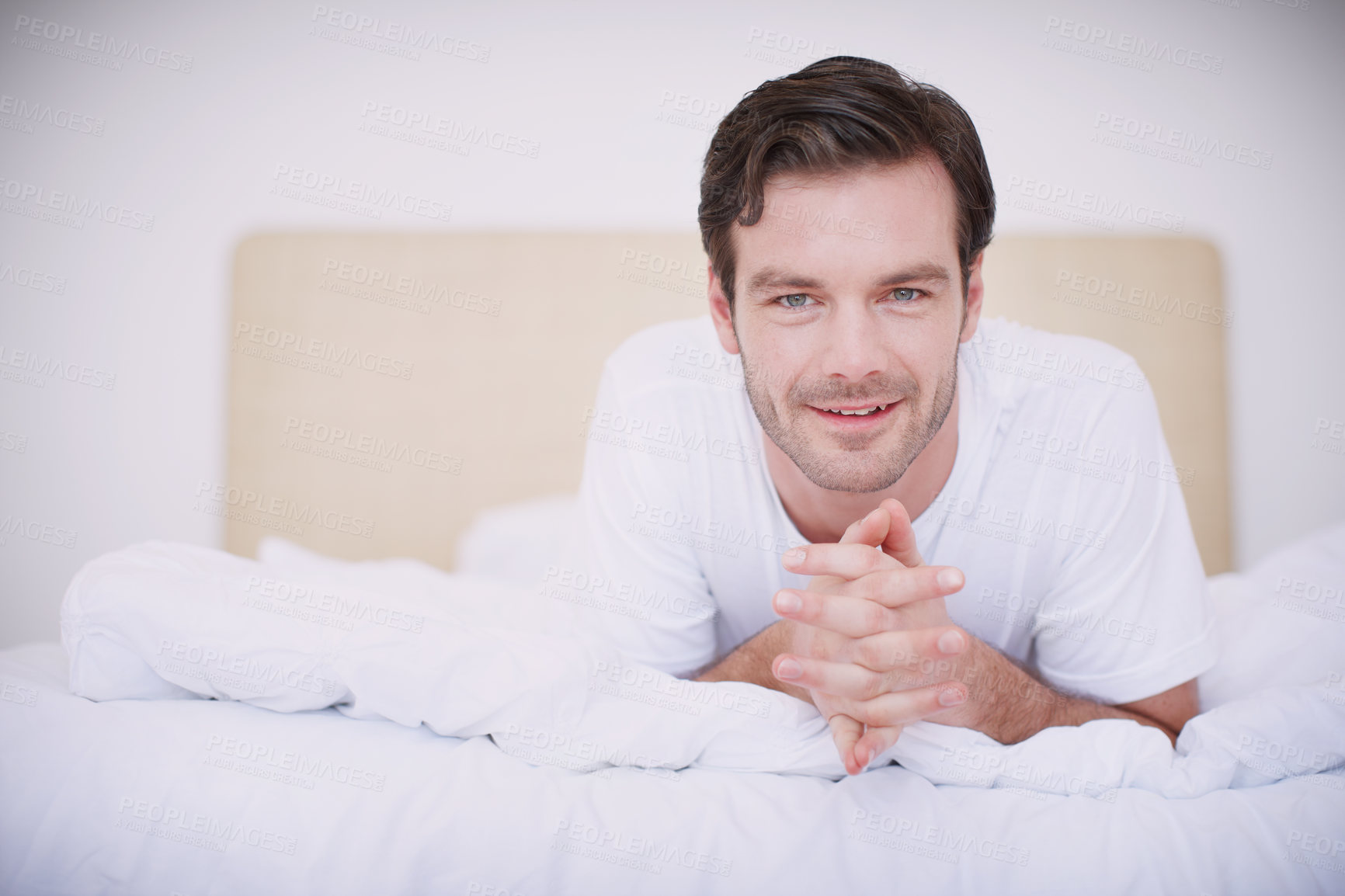 Buy stock photo Portrait of a handsome young man lying on his stomach on a bed with his hands clasped