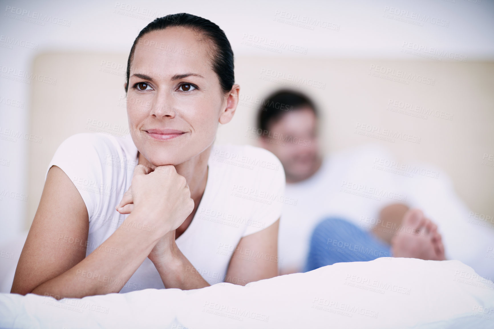 Buy stock photo A young woman looking thoughtful while lying on a bed with her husband in the background