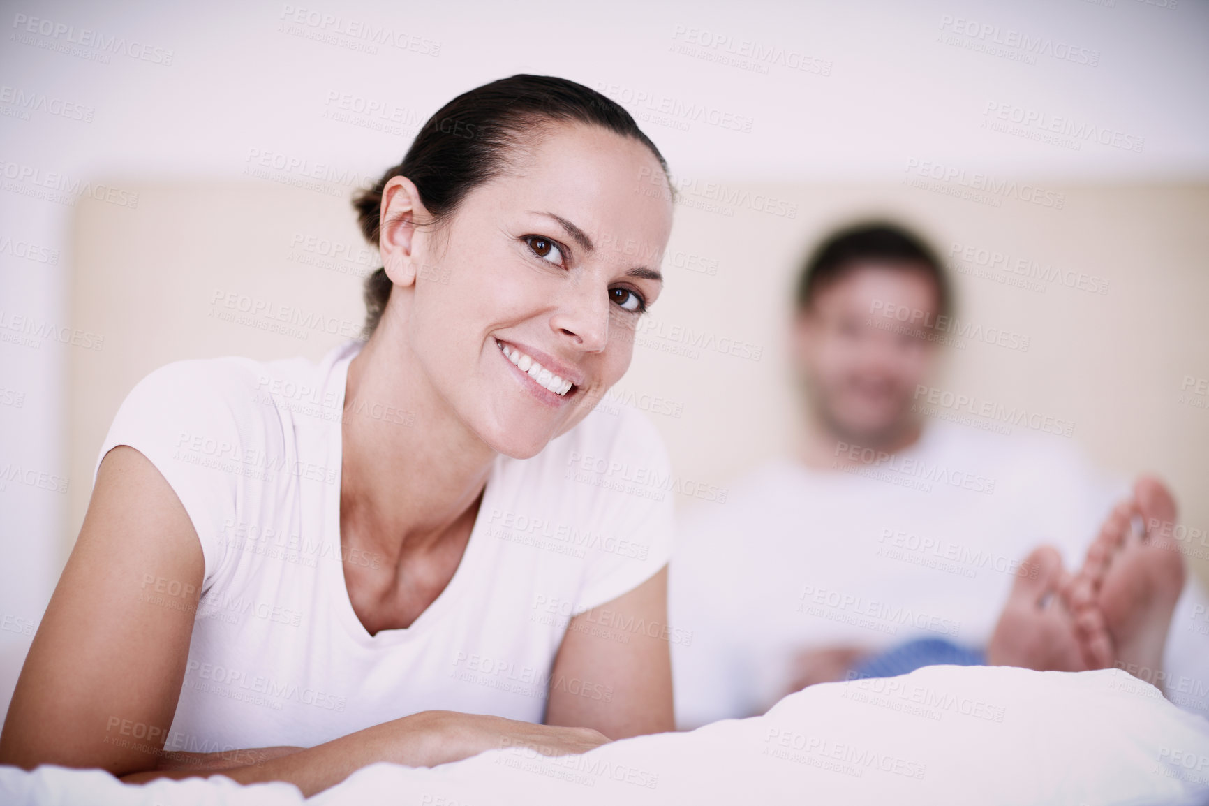 Buy stock photo A young woman smiling happily while lying on her bed with her husband in the background