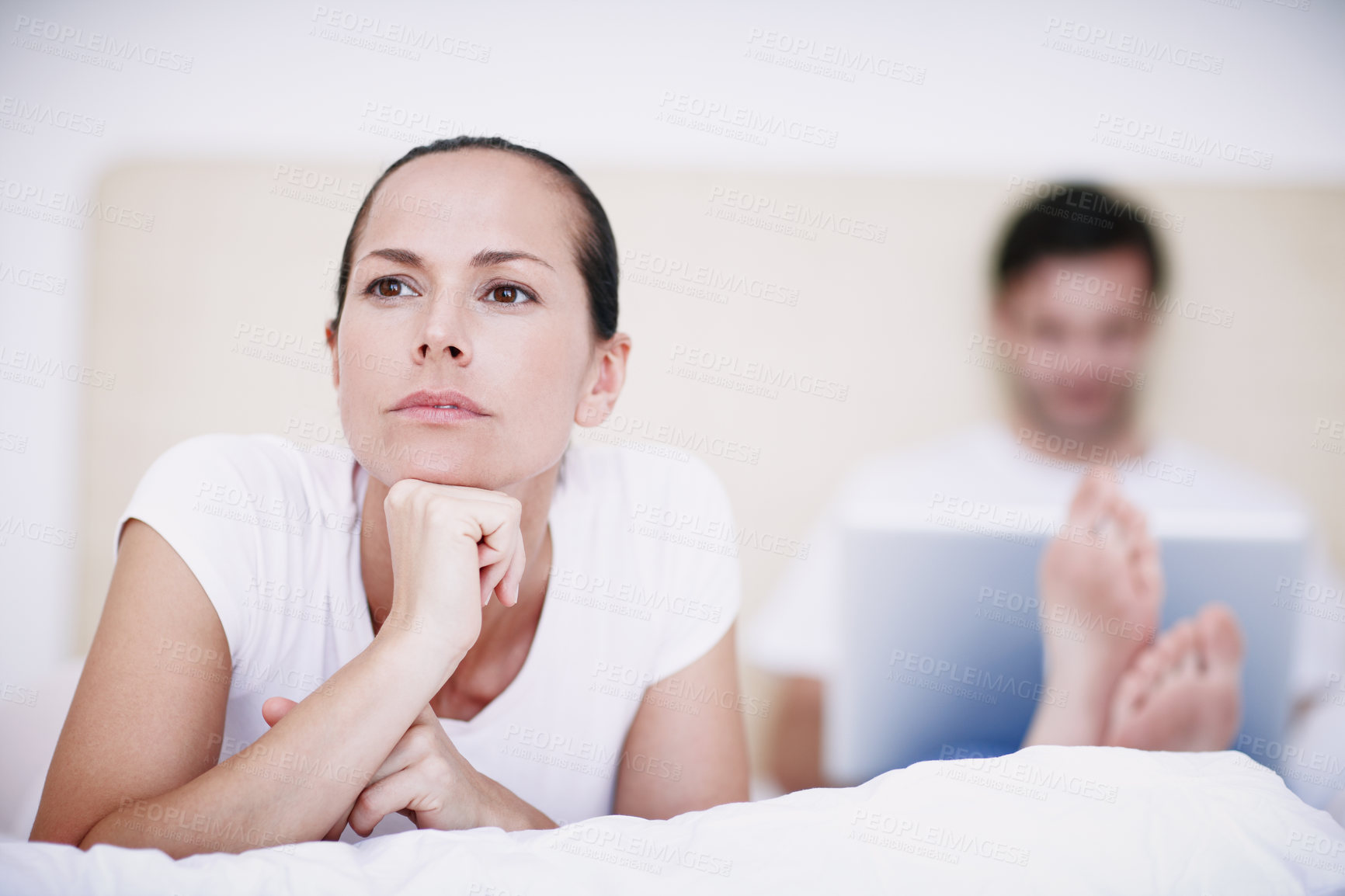 Buy stock photo A young woman looking displeased with her husband who is sitting in the background with his laptop