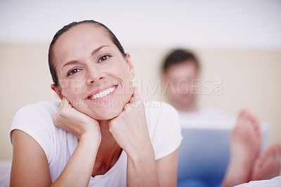 Buy stock photo Portrait of an attractive young woman lying on a bed while her husband works on a laptop in the background