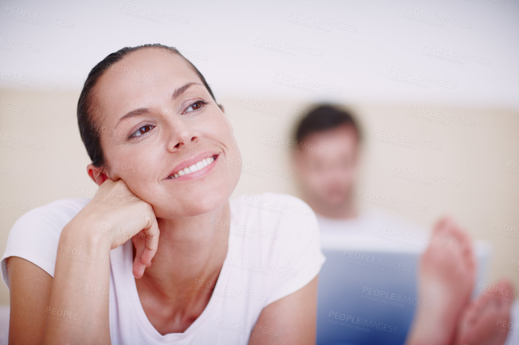 Buy stock photo A smiling young woman lying on a bed while her husband works on a laptop in the background