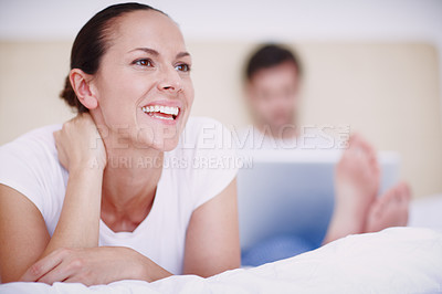 Buy stock photo A young woman lying on a bed while her husband works on a laptop in the background