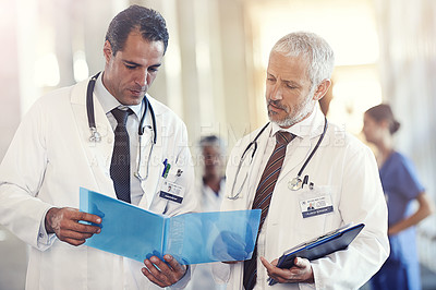 Buy stock photo Shot of two medical practitioners looking at a patient's chart with colleagues blurred in the background