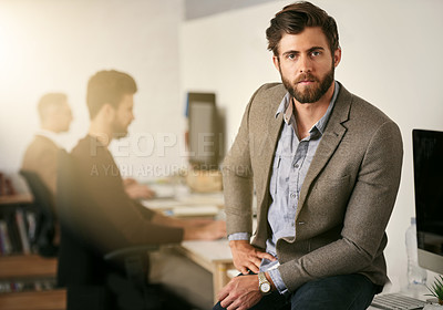 Buy stock photo Portrait of a young designer sitting on a desk with his colleagues blurred in the background