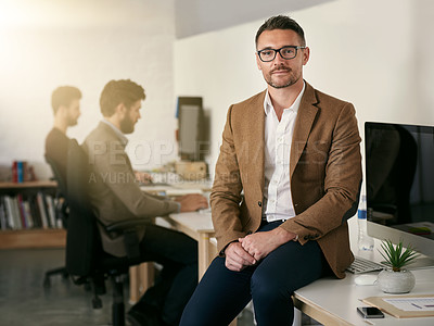 Buy stock photo Shot of a designer sitting on a desk with his colleagues blurred in the background