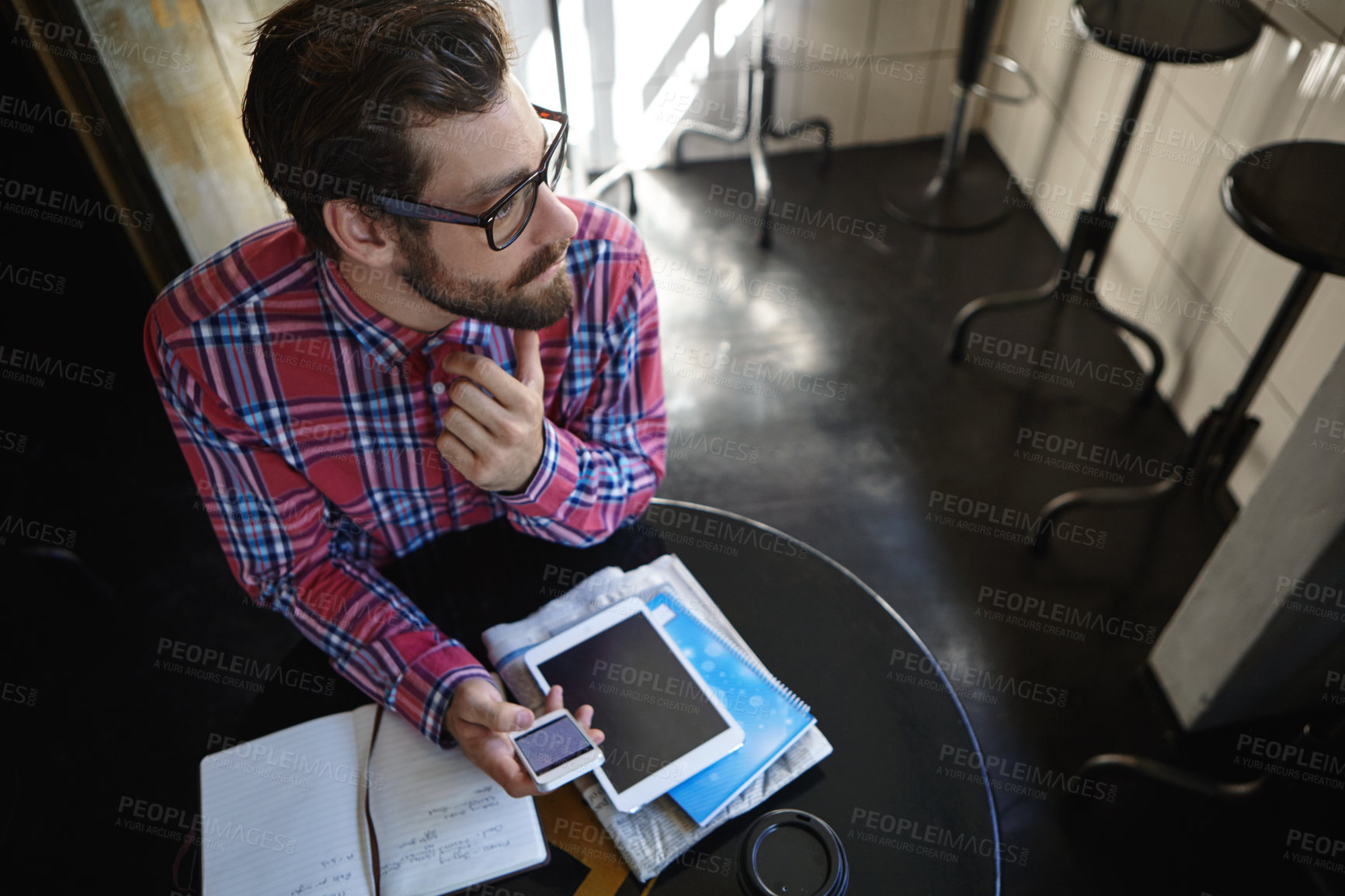 Buy stock photo Shot of a young man sitting at a table in a cafe with his cellphone, digital tablet and diary in front of him