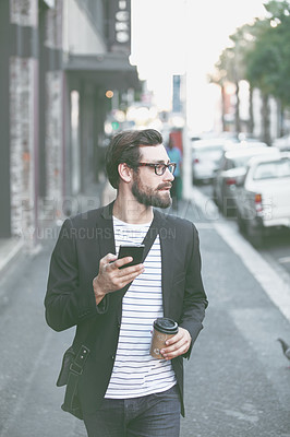 Buy stock photo A stylish young man carrying his cellphone and a takeaway coffee while walking in the city