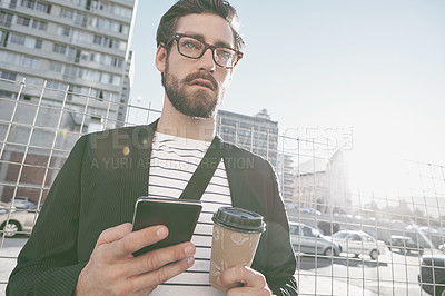 Buy stock photo A stylish young man holding his cellphone and a takeaway coffee in the city