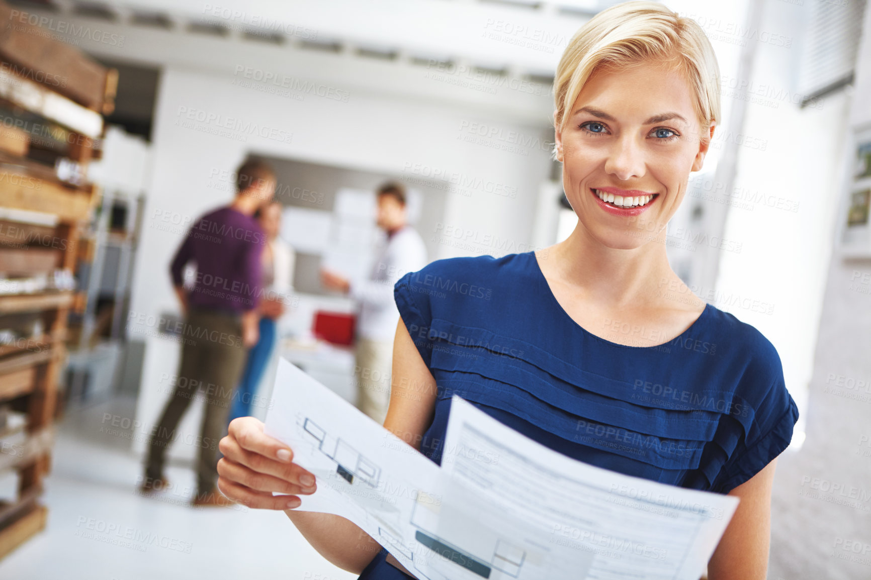 Buy stock photo Cropped portrait of a young female designer looking over some paperwork with her colleagues in the background